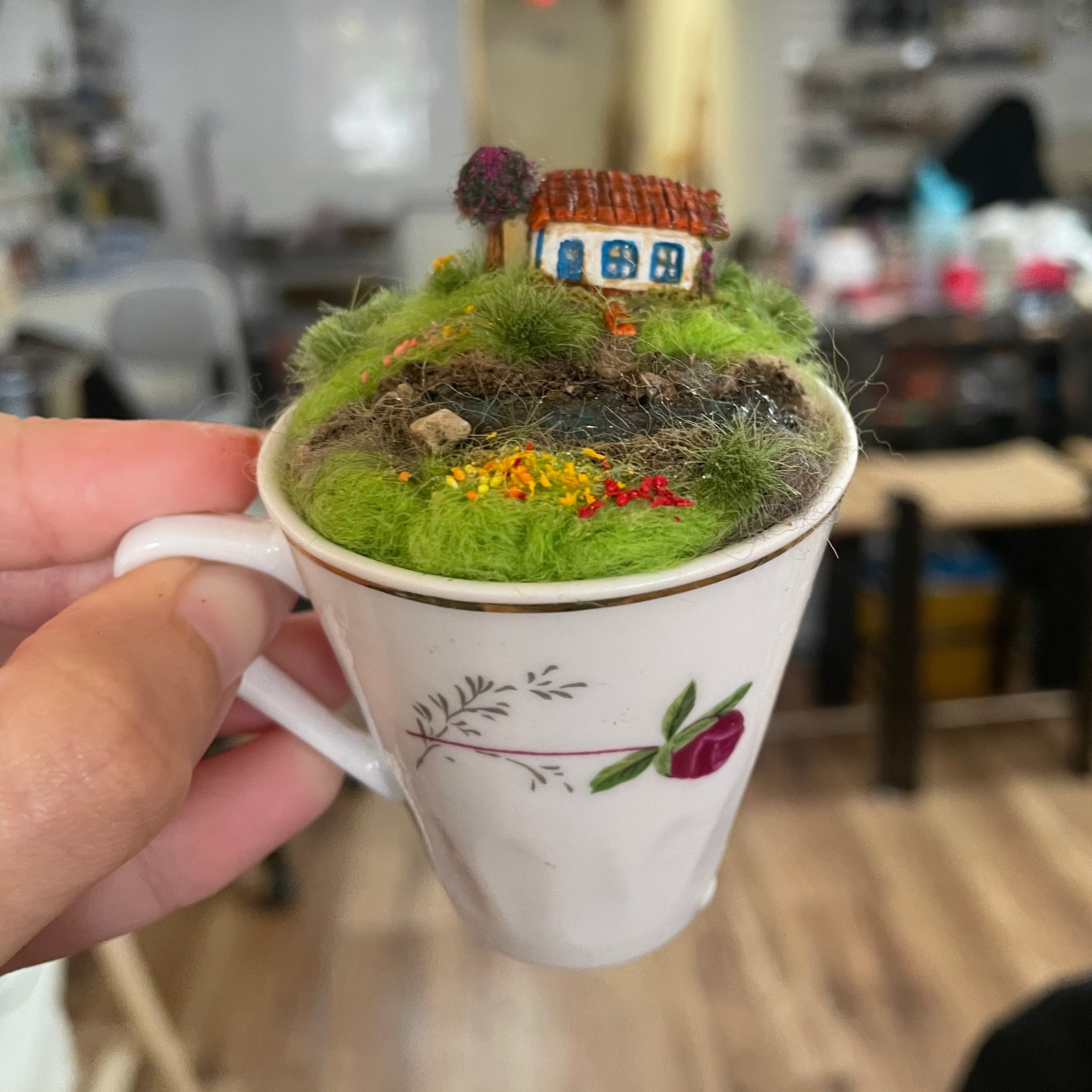 A hand holds a small miniature garden terrarium made from a white ceramic teacup, filled with artificial moss, tiny rocks, and a tiny house with a red roof in a real indoor room with blurred furniture and lights in the background.