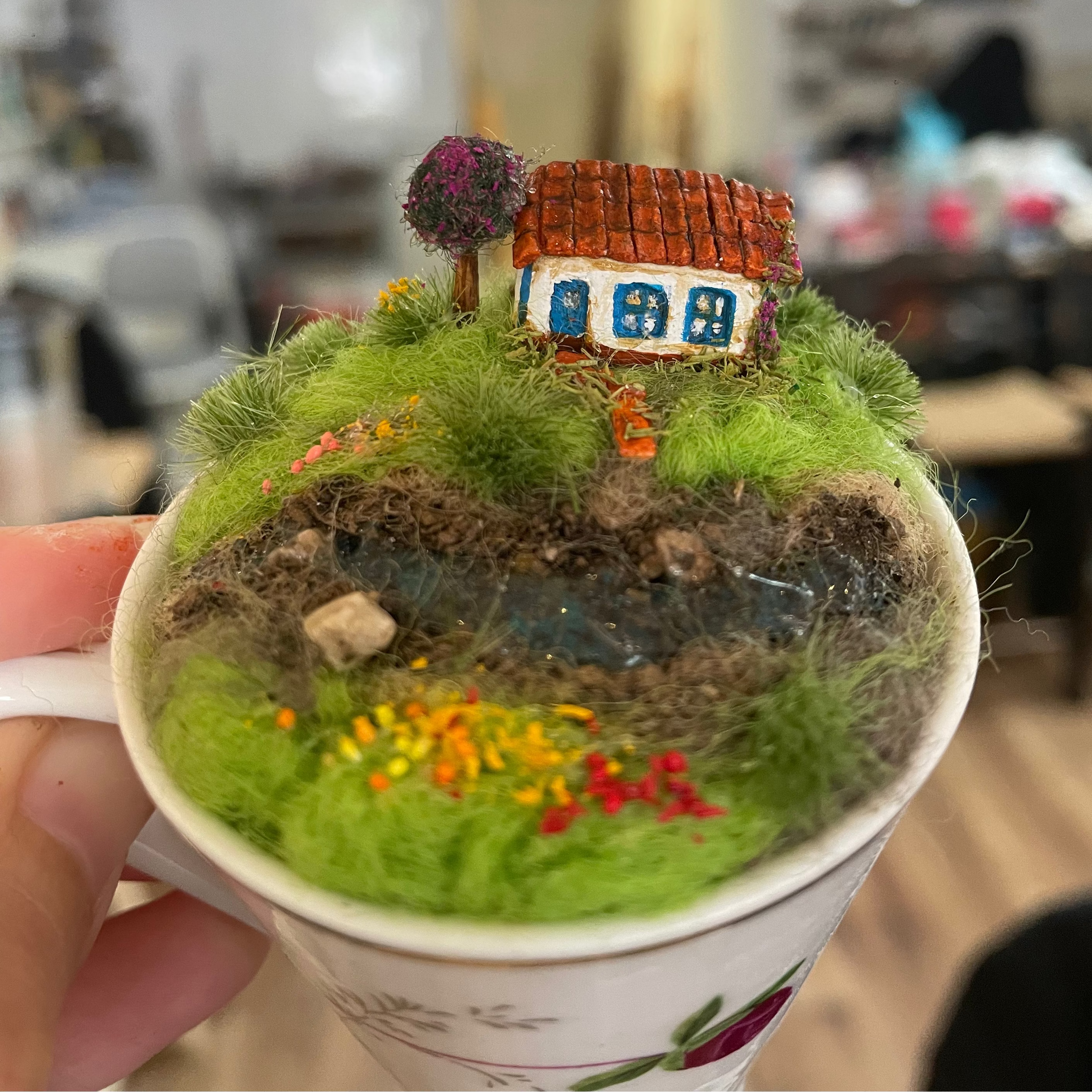 A miniature fairy garden terrarium made inside a white ceramic mug, featuring a tiny white house with a brown roof, green grass, and small colorful flowers, held by a hand in a blurred indoor setting.