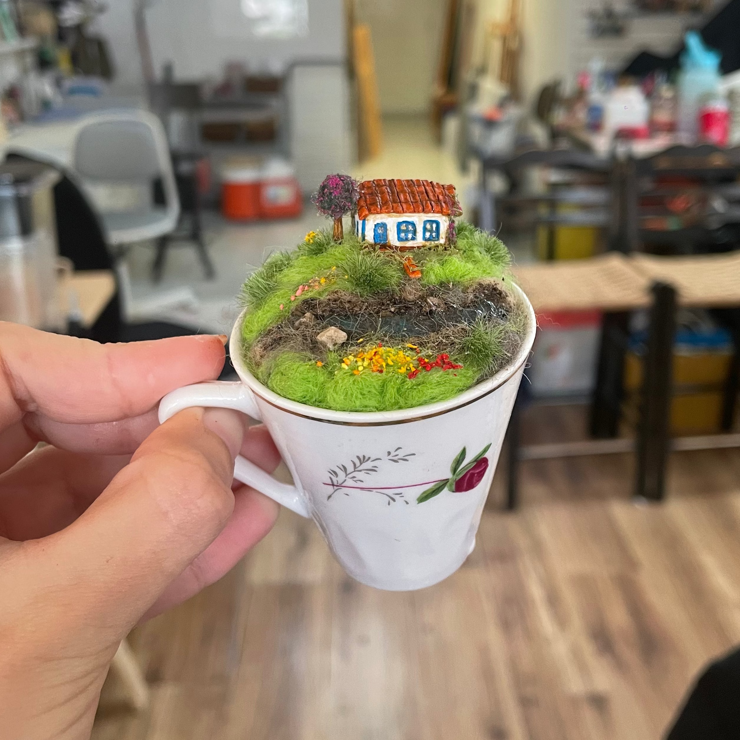A hand holds a miniature fairy garden inside a white ceramic teacup with a floral print, set against a blurred workshop or studio background.
