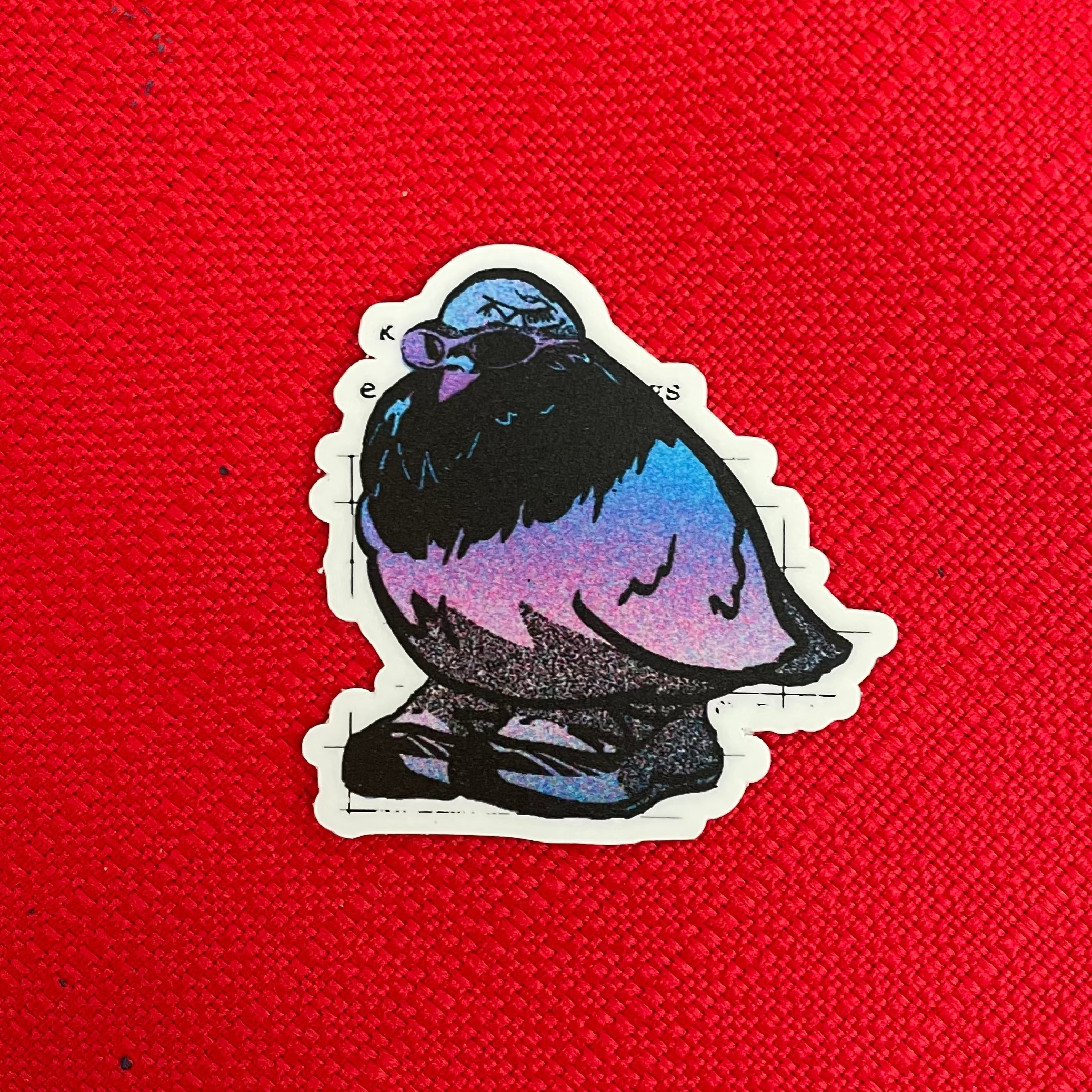 A die-cut sticker featuring a stylized pigeon with a blue, purple, and black gradient body, placed on a textured bright red fabric background.