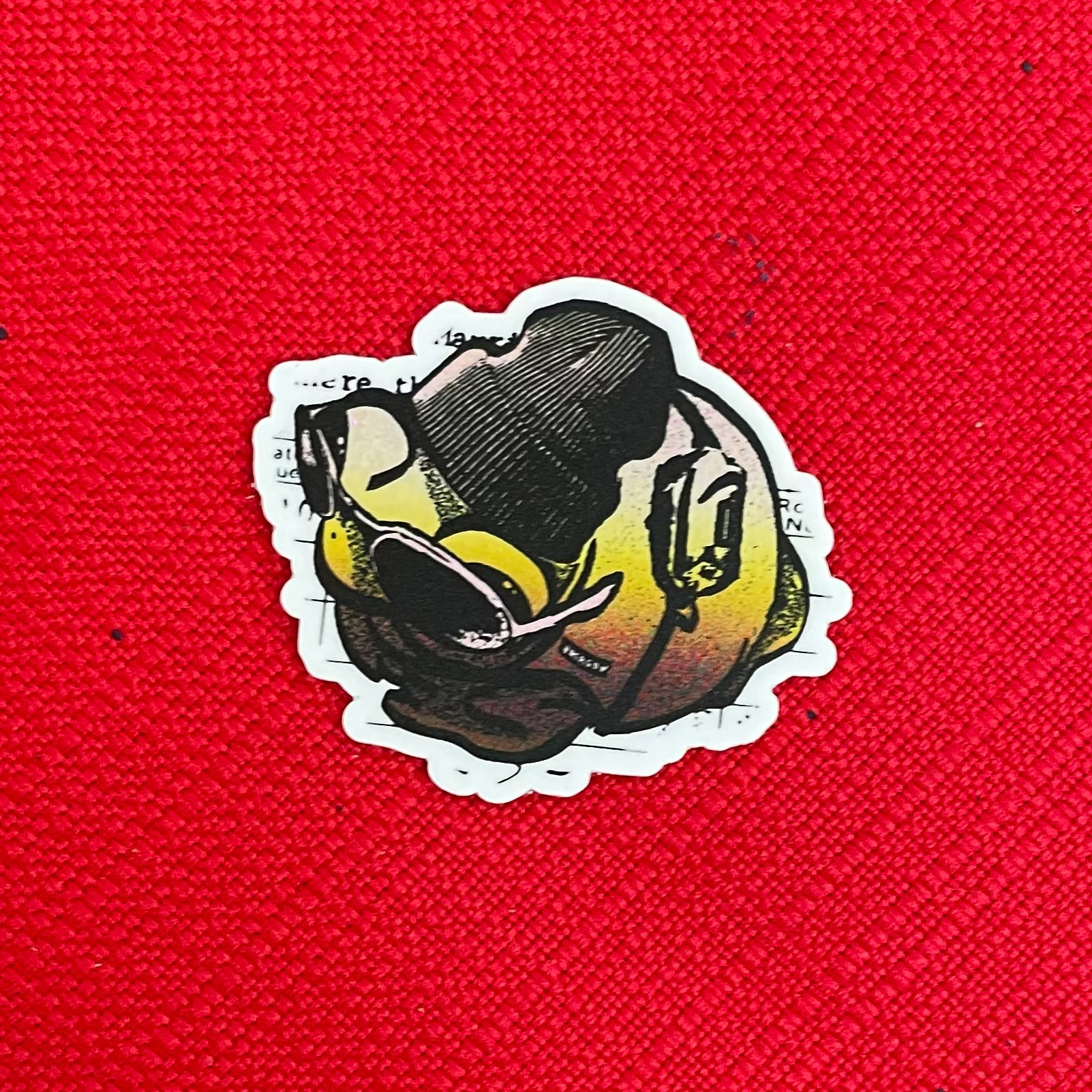 A small die-cut sticker featuring a stylized frog wearing Patagonia clothing, and a white outline, placed on a bright red textured fabric background.