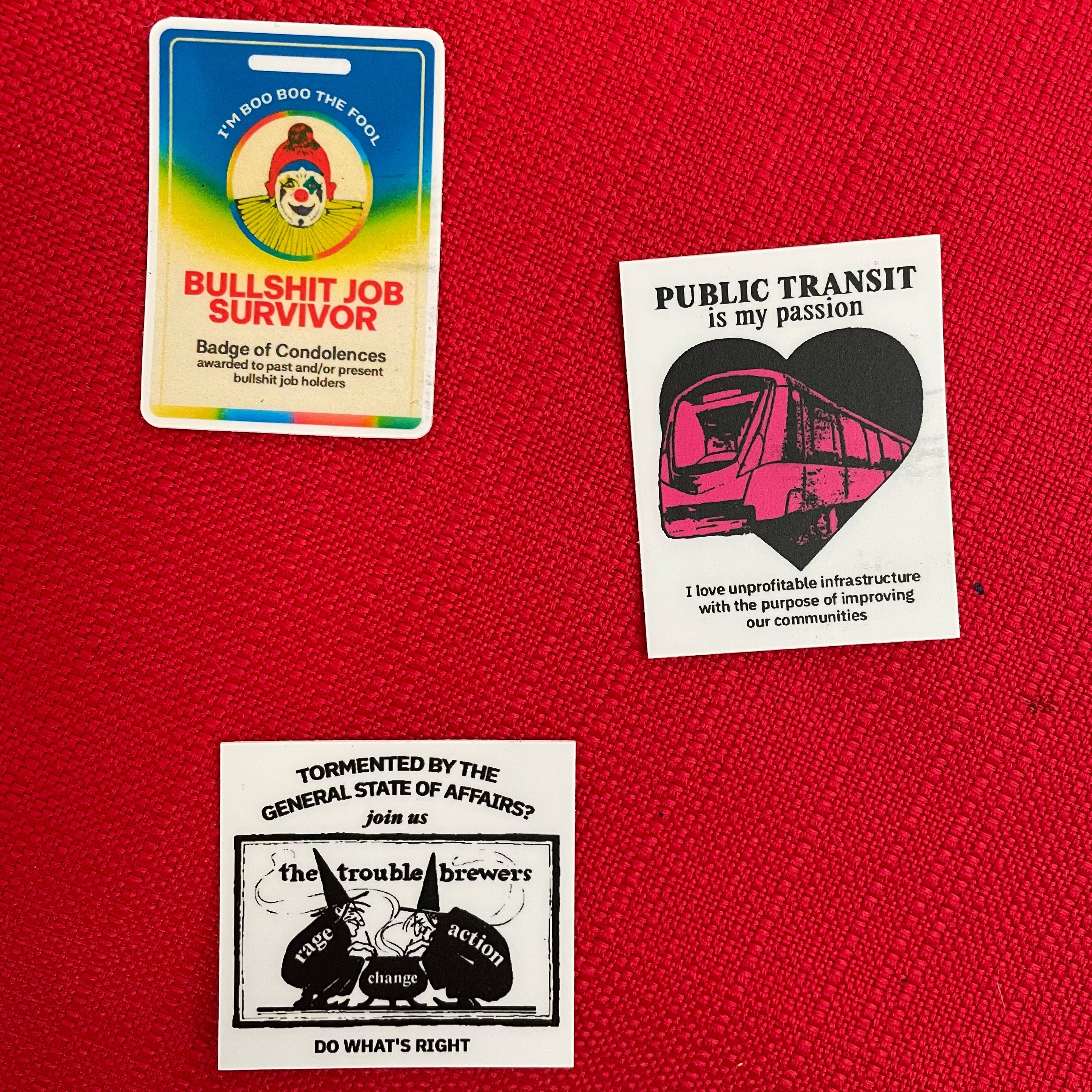Three novelty stickers displayed on a textured red fabric background. The stickers feature satirical and political designs: a 'Bullshit Job Survivor' badge, a transit worker pride sticker with a pink train on a black heart, and a 'Tormented by the General State of Affairs' sticker.