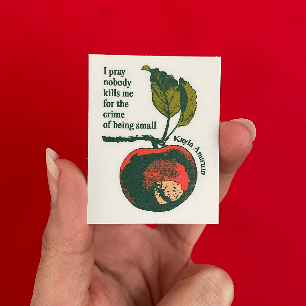 A hand holding a small white sticker featuring a red apple with a mouse inside and the quote, 'I pray nobody kills me for the crime of being small' by Kayla Ancrum, against a solid bright red background.