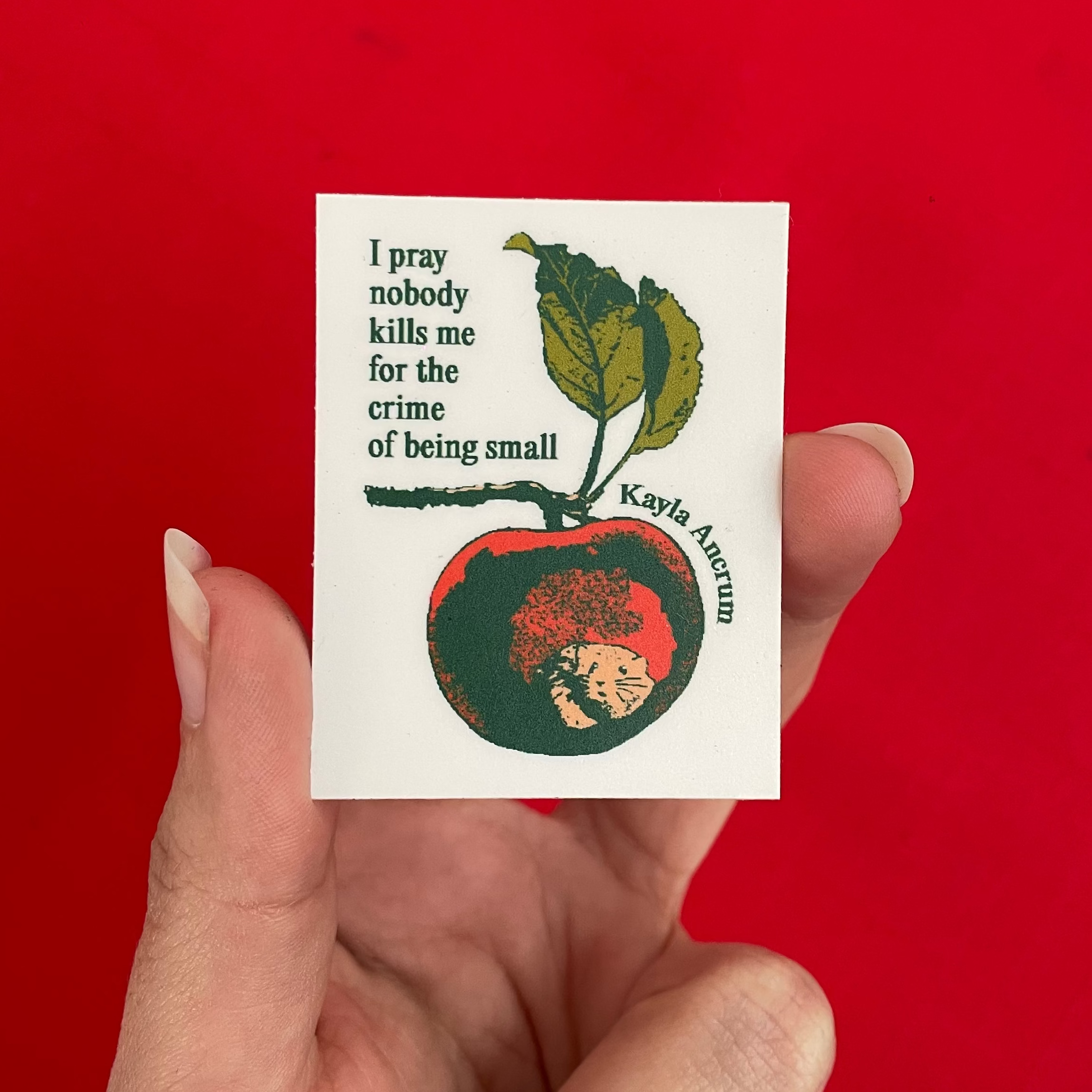 A hand holding a small white sticker featuring a red apple with a mouse inside and the quote, 'I pray nobody kills me for the crime of being small' by Kayla Ancrum, against a solid bright red background.