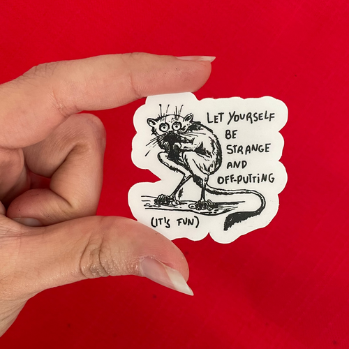A hand holds a die-cut sticker featuring a black ink sketch of a lemur-looking creature and the text 'LET YOURSELF BE STRANGE AND OFF-PUTTING (IT'S FUN)' against a solid bright red background.