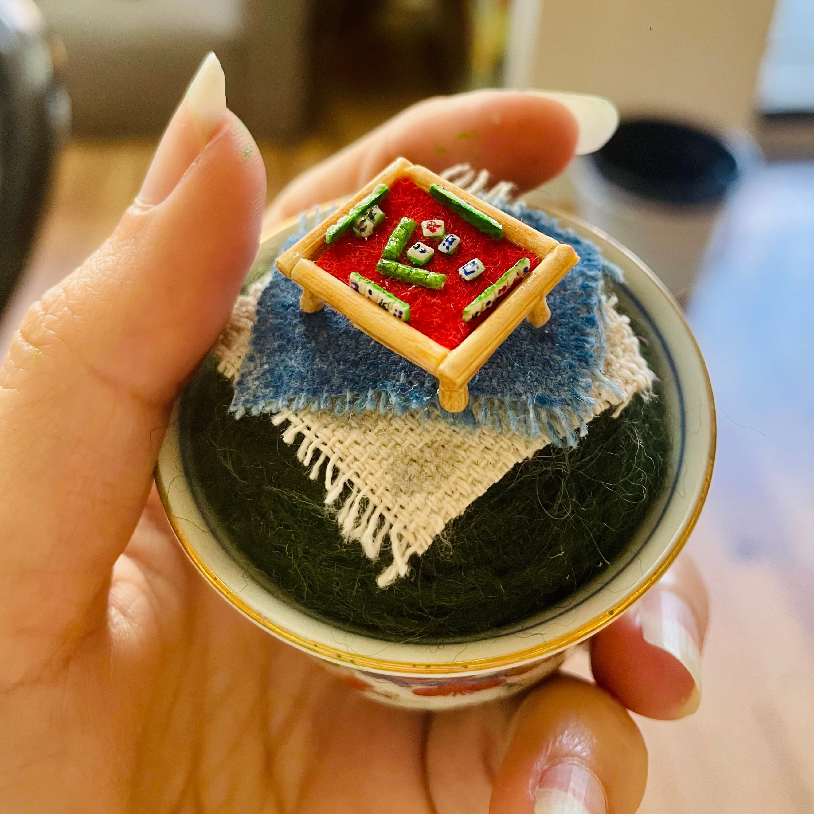 A hand holding a miniature mahjong set inside a small bowl. The set includes a red felt playing field, green tiles, white dice, a blue mat, and a beige woven runner, with a gold-rimmed bowl base.