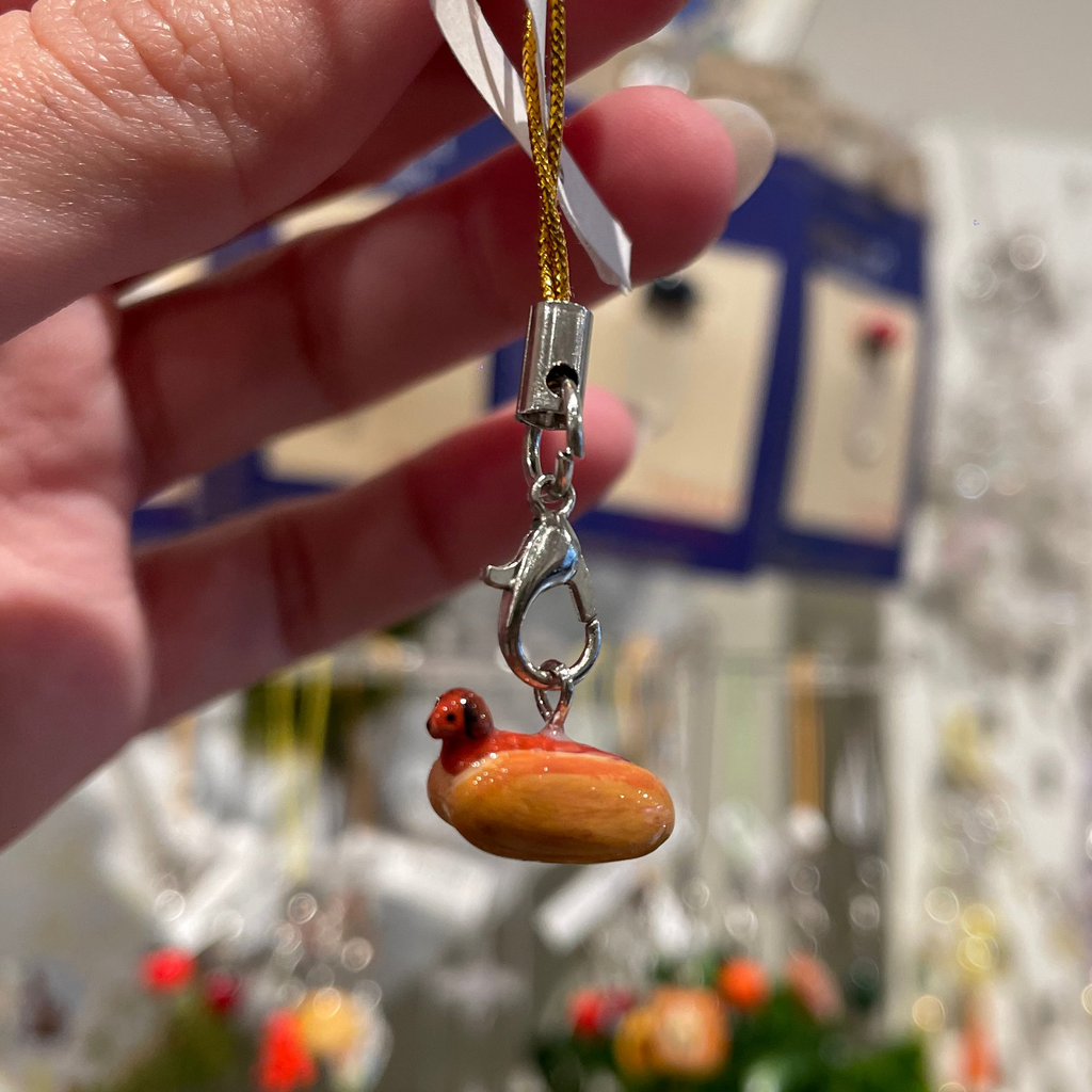 A hand holds a small charm of a hot dog, where the sausage is a tiny dog with its head poking out the bun, attached to a gold cord and silver clasp.