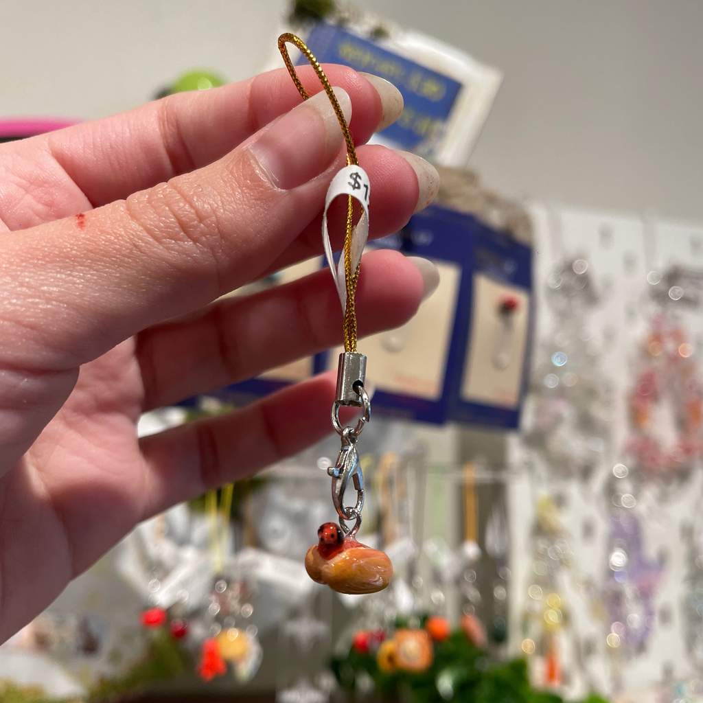 A hand holds a small charm of a hot dog, where the sausage is a tiny dog with its head poking out the bun, attached to a gold cord and silver clasp.