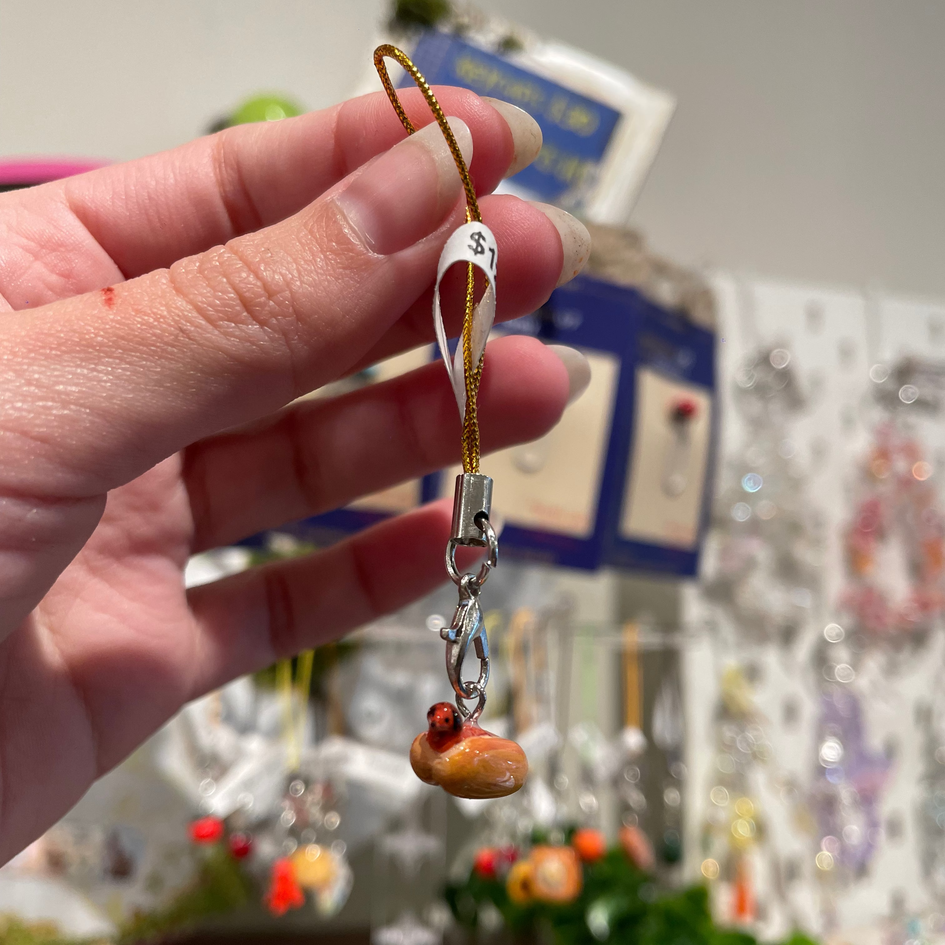 A hand holds a small charm of a hot dog, where the sausage is a tiny dog with its head poking out the bun, attached to a gold cord and silver clasp.