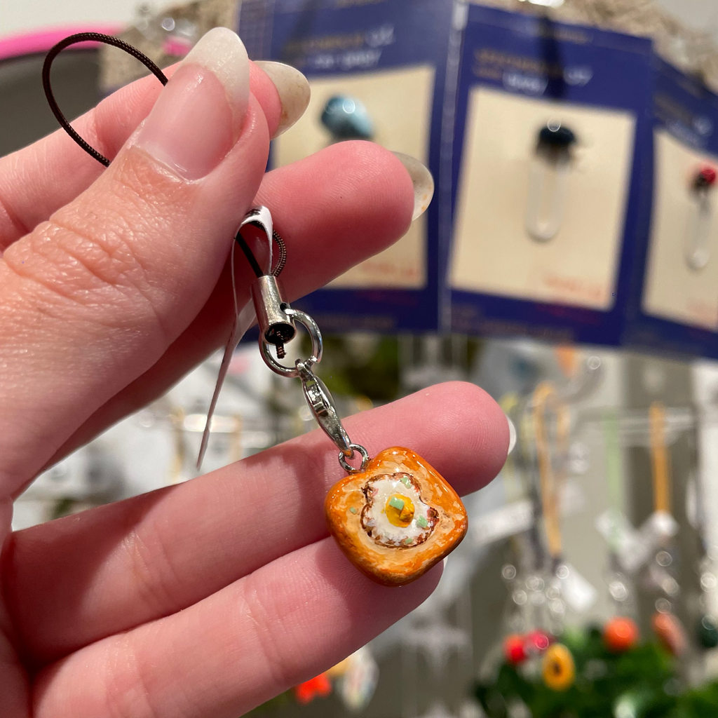 A hand holds a miniature fried egg on toast charm with a silver clasp.