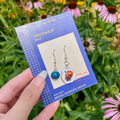 A hand holds a blue card displaying a pair of dangling earrings outdoors in front of green foliage and pink-and-white coneflowers. The earrings have silver hooks, and include a round blueberry and a plate of stacked blueberry pancakes.