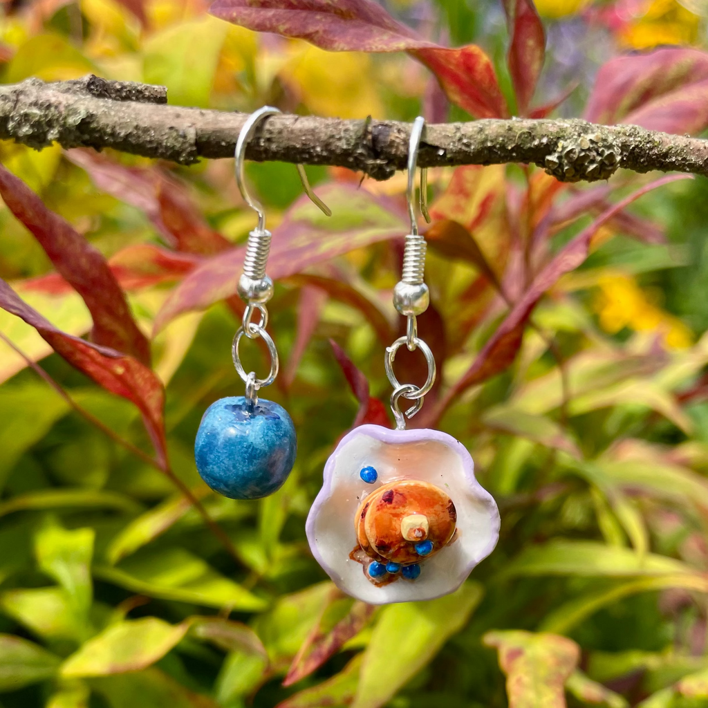 A pair of handmade dangle earrings hanging from a tree branch outdoors. One is a round blueberry, the other is a plate of stacked blueberry pancakes.
