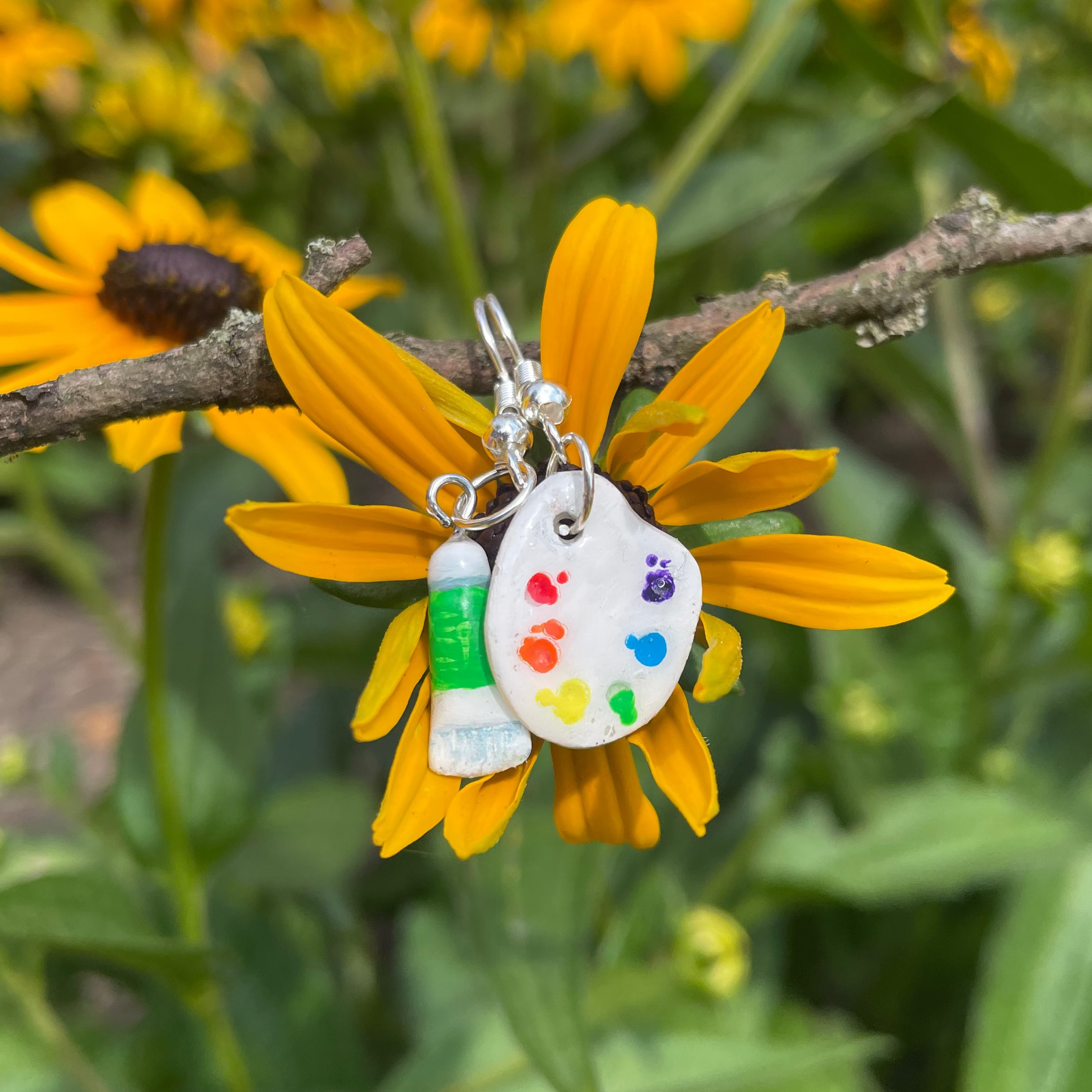 Small earrings featuring a white paint palette and paint tube design with colorful paint splatters, photographed against yellow flowers and green foliage.