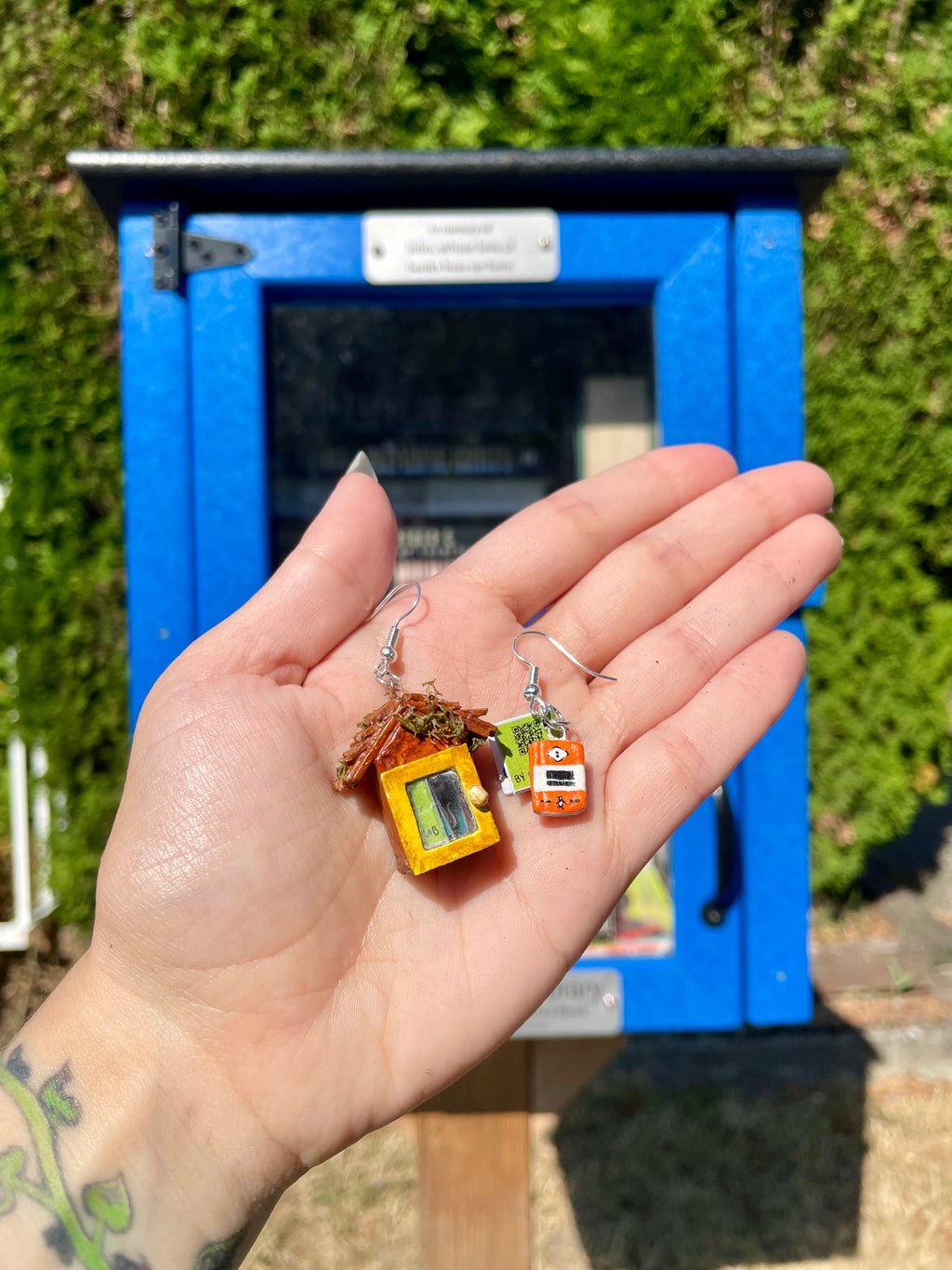 A hand holding a pair of miniature charm earrings in front of a bright blue kiosk with green foliage in the background.